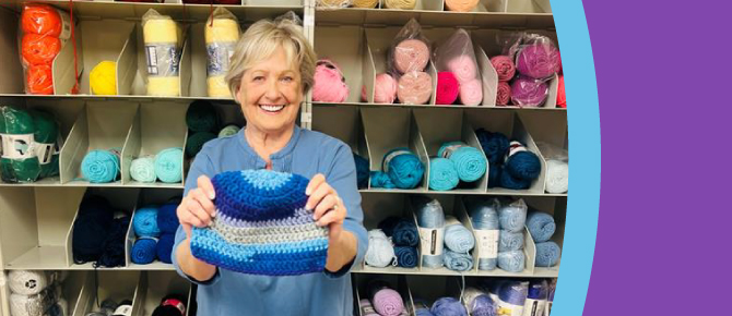 Kathy DeJoseph holding a knitted hat in front of wall of yarn